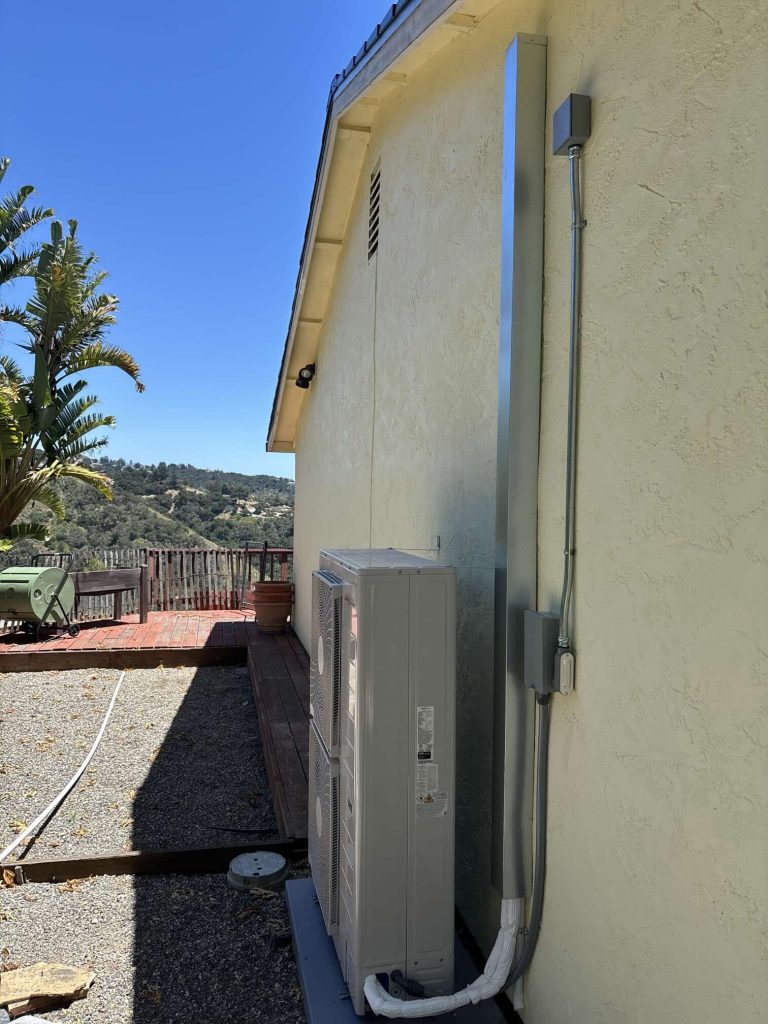 Slim heat pump condenser mounted against stucco wall at a Carmel Valley property with hillside backdrop, installed by Bellows